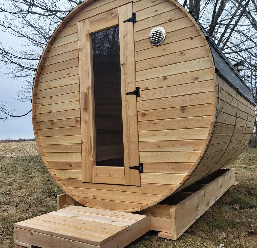 Cedar barrel sauna with a glass-panel door and a small front deck, set on a lawn at the edge of a field.