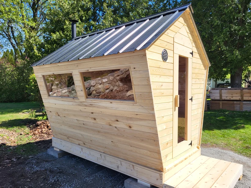 Cedar cabin sauna with a steep black metal roof, two long windows and a chimney, on a gravel pad in a backyard.