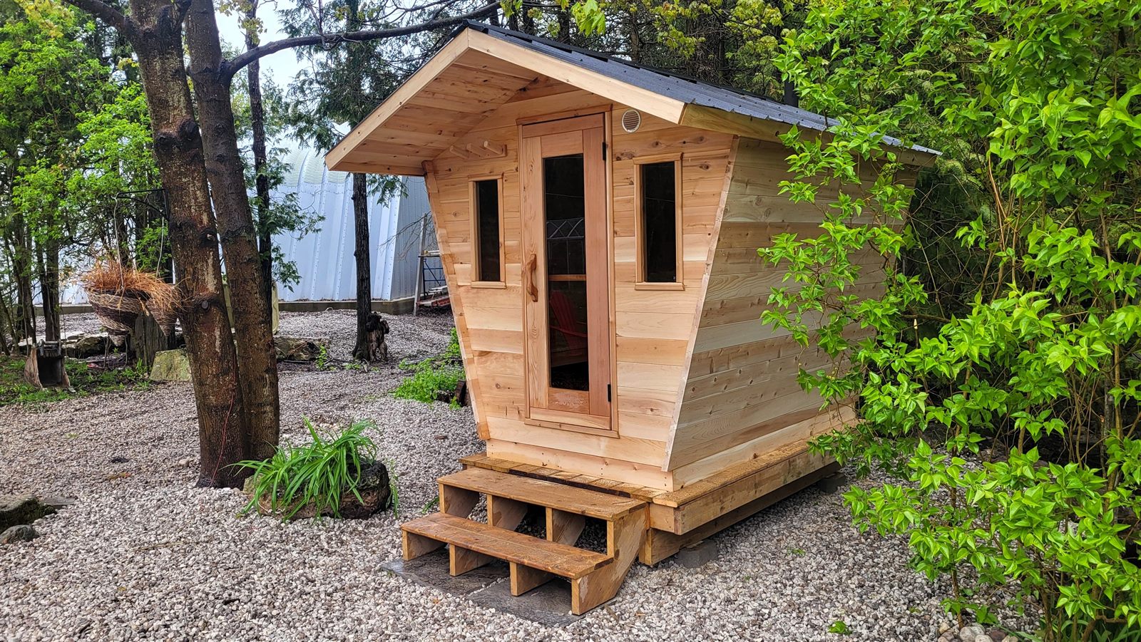 Small cedar cabin sauna with a flared roof and wooden steps, set on a gravel pad among trees in a backyard.