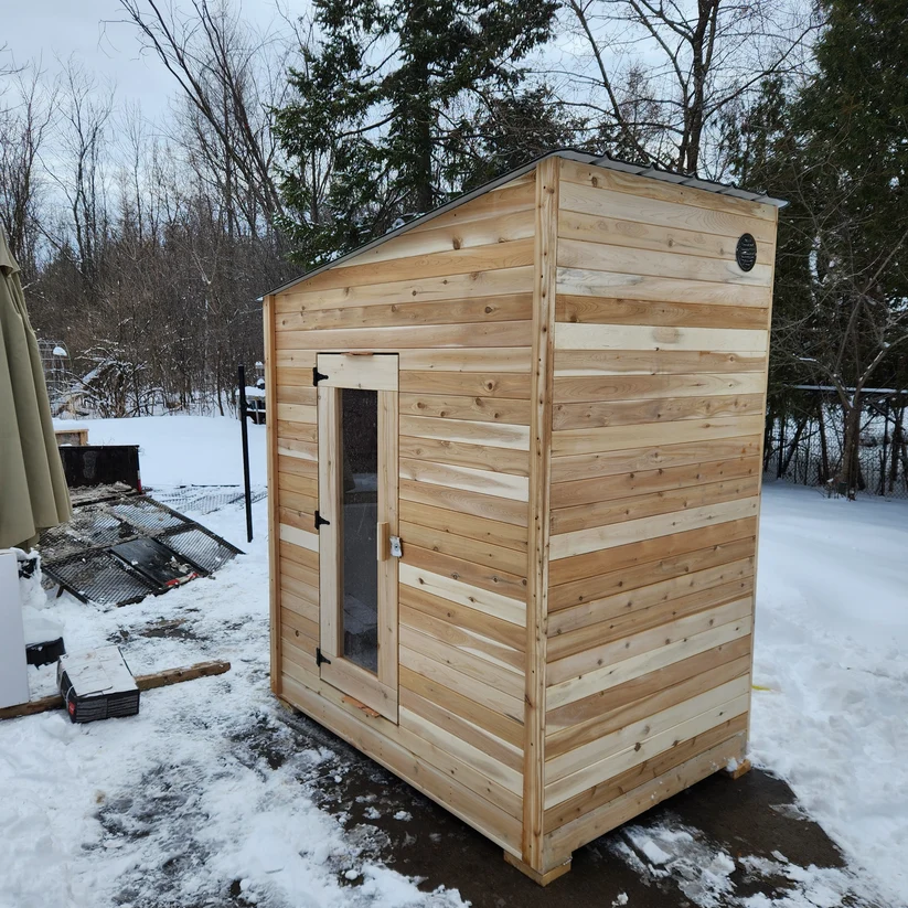 Cedar cabin sauna standing in a snow-covered yard on a grey winter day.