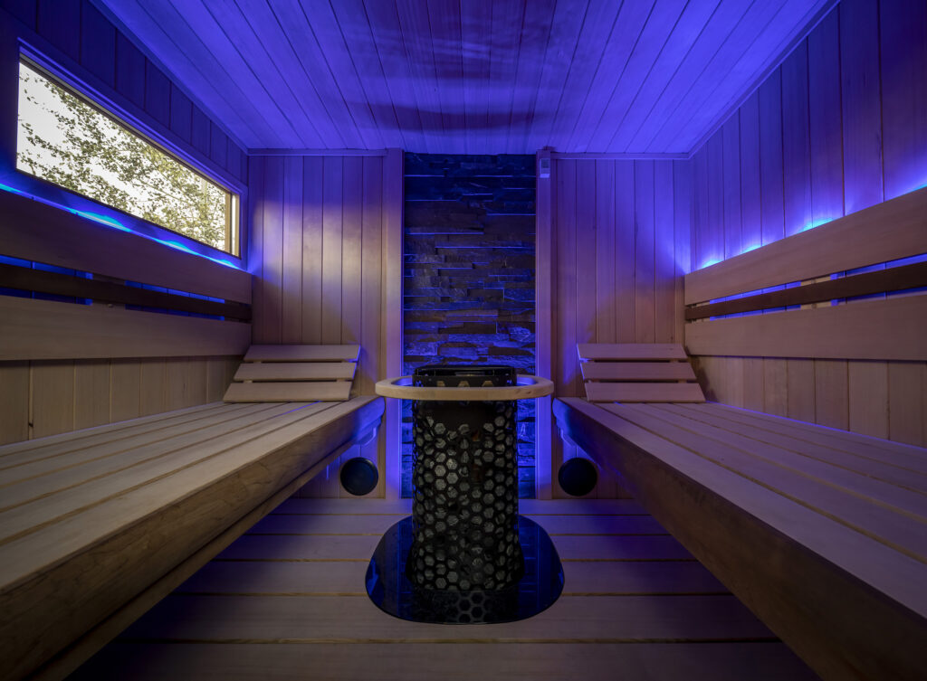 Indoor sauna interior lit in blue, with two facing cedar benches and a heater in a stone alcove.